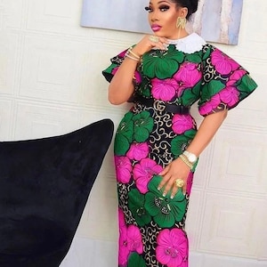 Puede incluir: Vestido largo con estampado floral de grandes flores verdes y rosas sobre fondo negro. Cuello blanco, mangas cortas y cinturón negro. El look se completa con joyas doradas y un reloj.