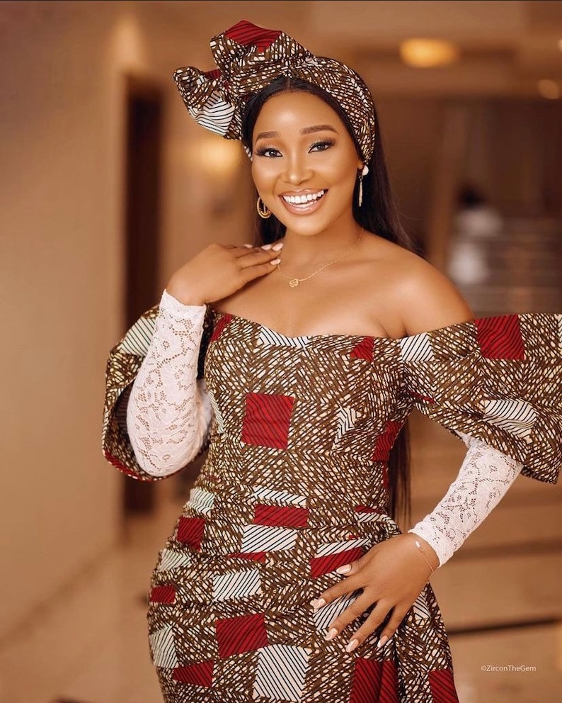 May include: An off-the-shoulder dress and matching head wrap. The dress features a geometric pattern in brown, red, and white. The sleeves are white lace. The woman is wearing gold earrings and a necklace.