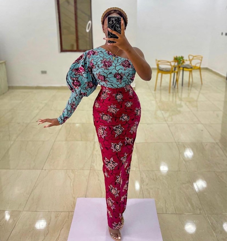 May include: A one-shoulder dress with a floral pattern. The top is light blue with pink flowers, and the skirt is a deep red with a similar floral design. The dress has a fitted silhouette and a long sleeve on the left arm.