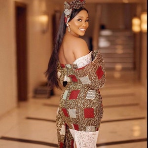 May include: A woman wearing a patterned dress with a matching head wrap. The dress features a brown, red, and white geometric design. The dress has a white lace trim at the bottom. The woman is smiling and looking over her shoulder.