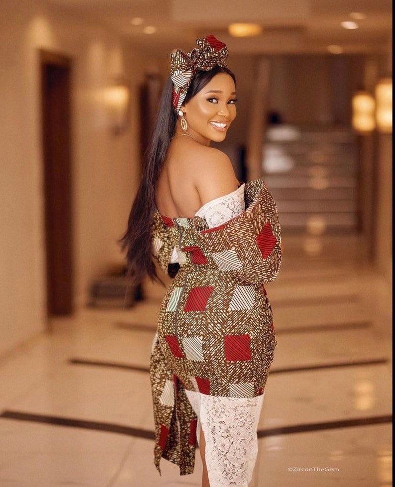 May include: A woman wearing a patterned dress with a matching head wrap. The dress features a brown, red, and white geometric design. The dress has a white lace trim at the bottom. The woman is smiling and looking over her shoulder.