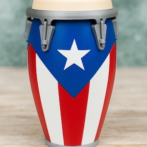 May include: A conga drum featuring the flag of Puerto Rico. The drum has a blue top with a white star, red and white stripes, a beige drumhead, and gray hardware. The drum is cylindrical with a silver base.