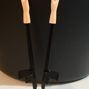 Puede incluir: Un utensilio de servir negro con dos brazos, cada uno terminando en una mano de tono carne pálido. El utensilio está diseñado para sujetar o levantar objetos, con una base en forma de U para mayor estabilidad. El fondo es oscuro.