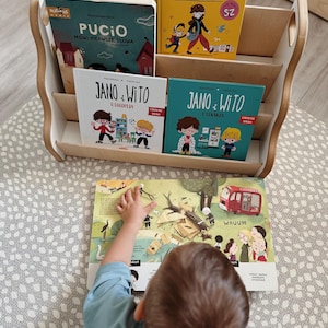 May include: A light-colored wooden bookshelf holding four children's books titled Jano i Wito and Pucio. An open illustrated book lies in front of the shelf, featuring a red vehicle, a fallen tree, and various characters in a colorful outdoor scene.