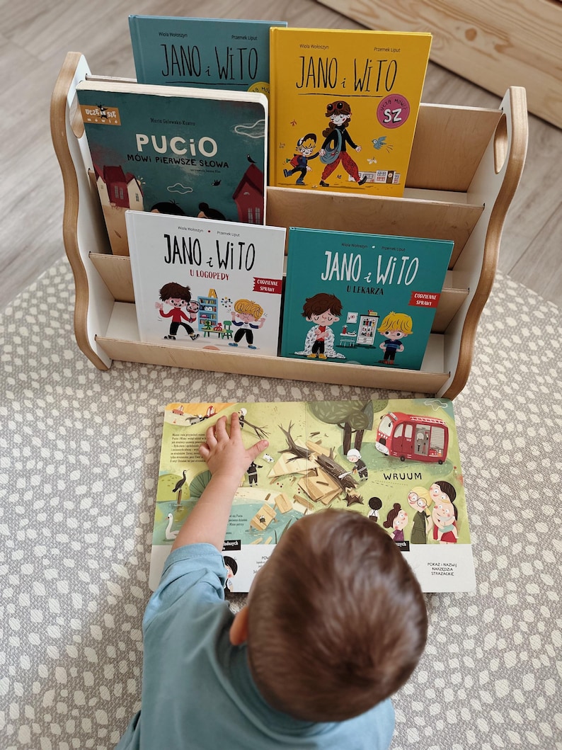 May include: A light-colored wooden bookshelf holding four children's books titled Jano i Wito and Pucio. An open illustrated book lies in front of the shelf, featuring a red vehicle, a fallen tree, and various characters in a colorful outdoor scene.