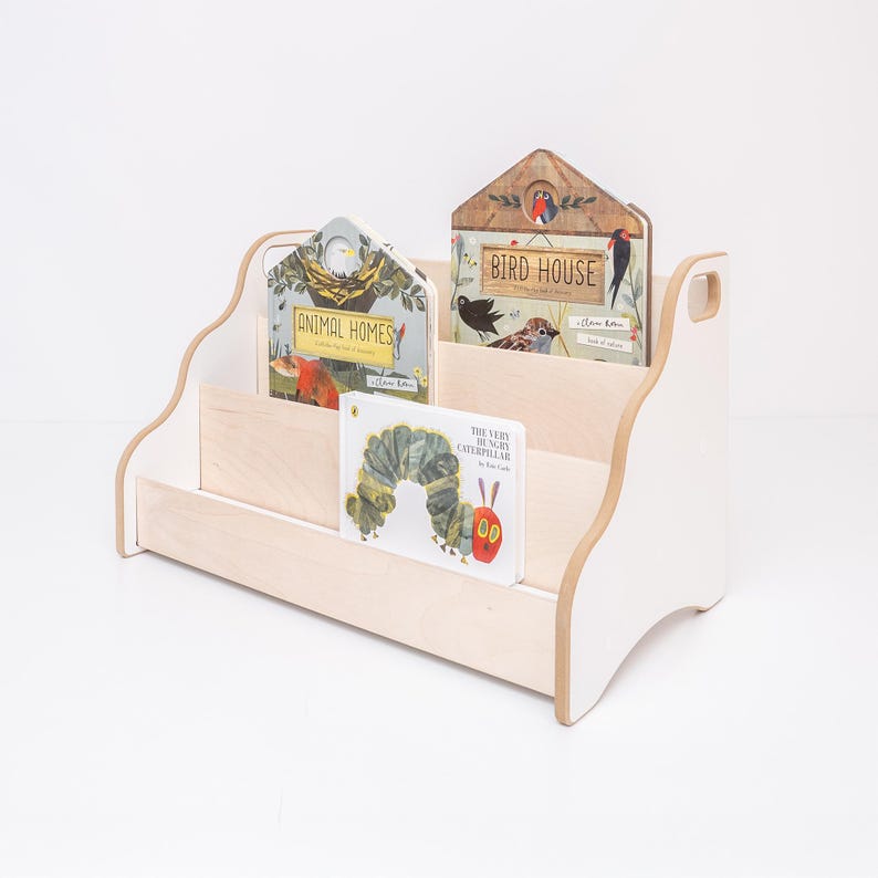 May include: A light-colored wooden bookshelf with two tiers and wavy side panels. The shelf holds three books: Animal Homes, Bird House, and The Very Hungry Caterpillar. The structure features a natural wood grain finish with white accents.
