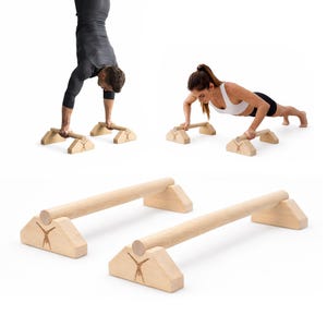 Könnte beinhalten: Ein Paar helle Holz-Parallettes mit zylindrischen Stangen, die auf zwei dreieckigen Sockeln ruhen. Jeder Sockel ist mit einer kleinen Gravur einer Person im Handstand versehen, die Stabilität für verschiedene Fitnessübungen bietet.