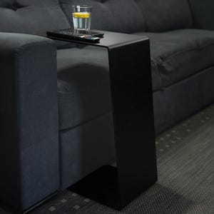 Pode incluir: Uma mesa lateral preta em forma de C colocada ao lado de um sofá cinza escuro. Um copo de água com limão e um controle remoto estão sobre a mesa. A mesa é de metal e tem um design moderno.