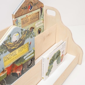 May include: A light-colored wooden bookshelf with two tiers and side handles. It holds three children's books: Bird House, Animal Homes, and The Very Hungry Caterpillar. The books feature colorful illustrations of birds, a fox, and a green caterpillar.