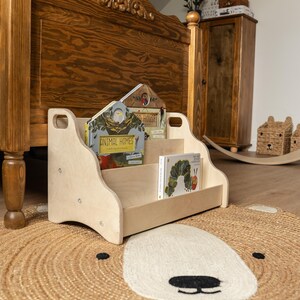 May include: A light-colored wooden bookshelf with two tiers and cut-out handles on the sides. The shelf holds two books, one titled Animal Homes and another featuring a caterpillar illustration, resting on a round woven rug with a bear face design.