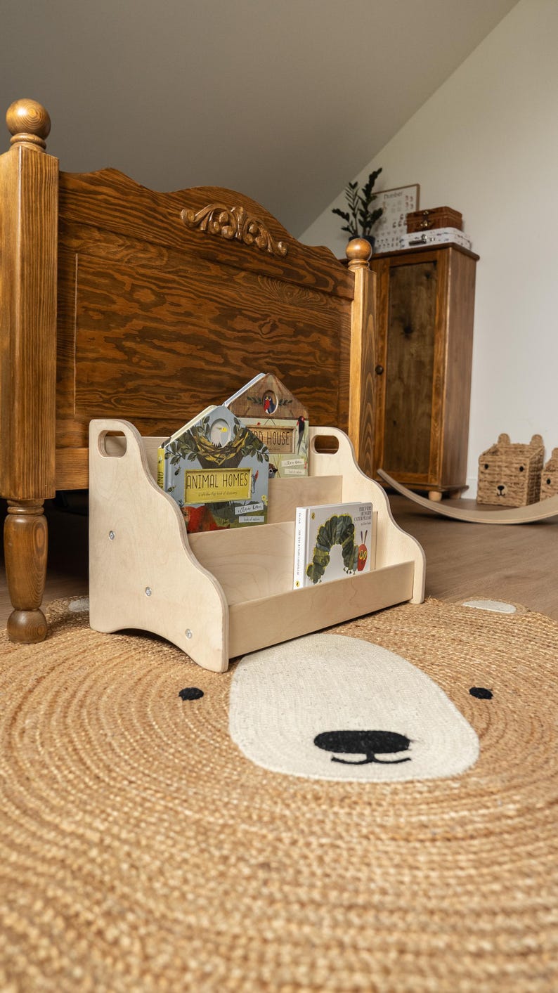 May include: A light-colored wooden bookshelf with two tiers and cut-out handles on the sides. The shelf holds two books, one titled Animal Homes and another featuring a caterpillar illustration, resting on a round woven rug with a bear face design.