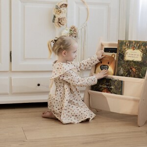 K&ouml;nnte beinhalten: Ein junges M&auml;dchen in einem Blumenkleid greift nach einem Buch in einem B&uuml;cherregal aus Holz. Das B&uuml;cherregal hat drei Regalbretter und ist mit bunten B&uuml;chern gef&uuml;llt. Das B&uuml;cherregal ist hellbraun und hat ein schlichtes Design.