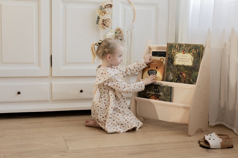 K&ouml;nnte beinhalten: Ein junges M&auml;dchen in einem Blumenkleid greift nach einem Buch in einem B&uuml;cherregal aus Holz. Das B&uuml;cherregal hat drei Regalbretter und ist mit bunten B&uuml;chern gef&uuml;llt. Das B&uuml;cherregal ist hellbraun und hat ein schlichtes Design.