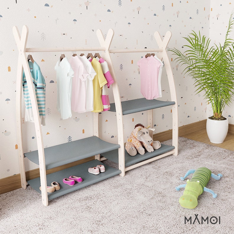 May include: White wooden clothing rack with two shelves and a hanging bar. The rack has a gray fabric shelf and a gray fabric bottom shelf. The rack is decorated with a stuffed giraffe and a stuffed green alligator. The rack is against a white wall with a pattern of small shapes.