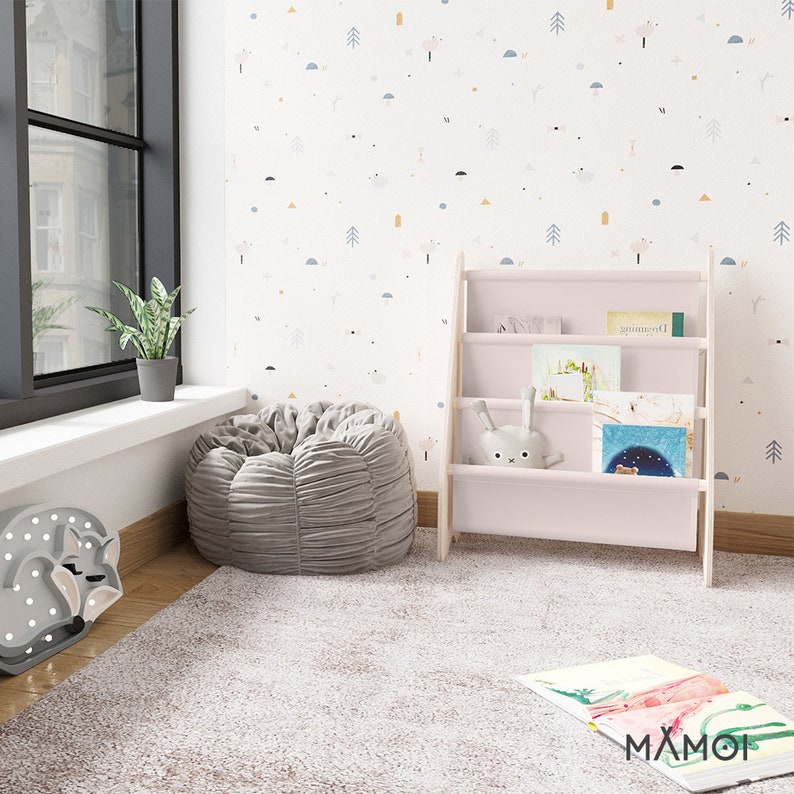 K&ouml;nnte beinhalten: Ein rosa Holz-Regal mit vier Regalbrettern, gef&uuml;llt mit Kinderb&uuml;chern. Ein pl&uuml;schiger grauer Sitzsack steht vor dem Regal, und ein wei&szlig;er Teppich bedeckt den Boden. Die Wand hinter dem Regal ist mit einer wei&szlig;en Tapete bedeckt, die ein sich wiederholendes Muster aus kleinen blauen, gelben und rosa geometrischen Formen aufweist.