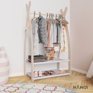 May include: A wooden clothing rack with two shelves and a hanging bar. The rack is painted white and has a light wood finish. The shelves are covered in a light gray fabric. The rack is decorated with clothes, shoes, and a wire basket filled with toys.