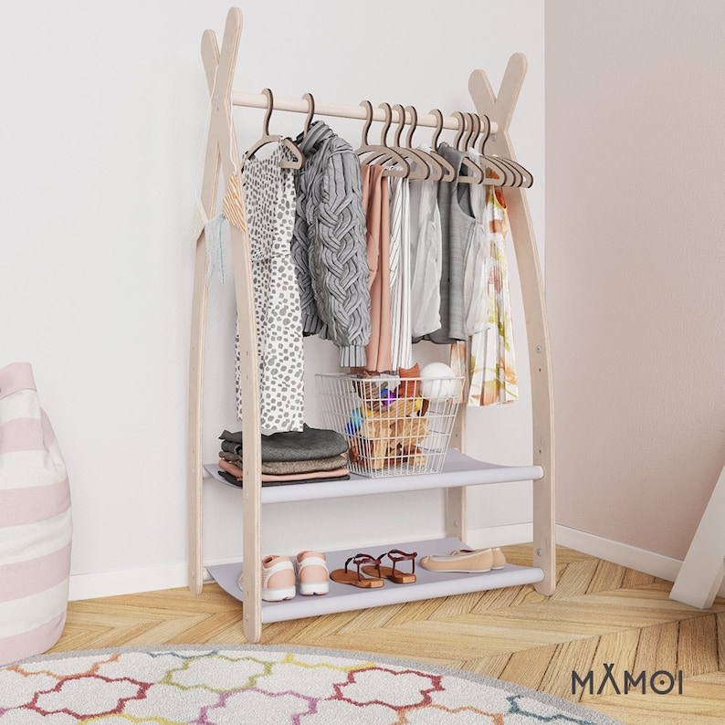 May include: A wooden clothing rack with two shelves and a hanging bar. The rack is painted white and has a light wood finish. The shelves are covered in a light gray fabric. The rack is decorated with clothes, shoes, and a wire basket filled with toys.
