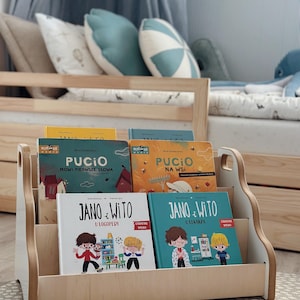 May include: A light-colored wooden bookshelf holding four children's books titled Pucio M&oacute;wi Pierwsze Słowa, Pucio Na Wsi, Jano i Wito U Logopedy, and Jano i Wito U Lekarza. The shelf has a low, open-front design with side cutouts for easy handling.