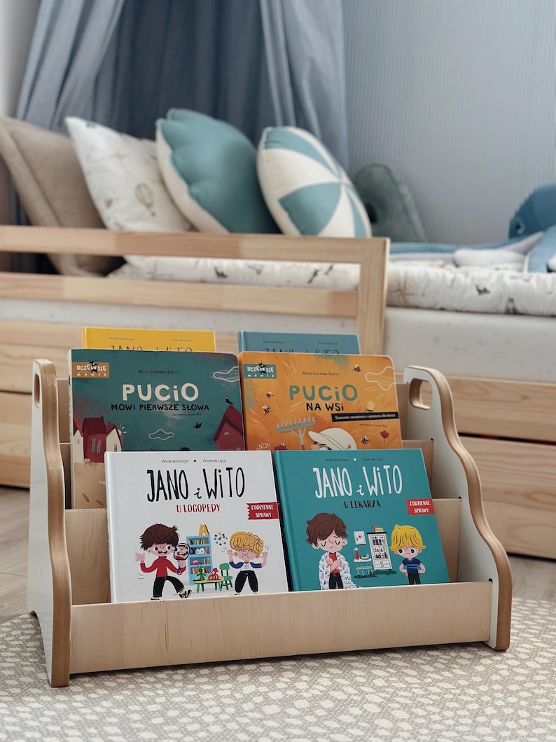 May include: A light-colored wooden bookshelf holding four children's books titled Pucio M&oacute;wi Pierwsze Słowa, Pucio Na Wsi, Jano i Wito U Logopedy, and Jano i Wito U Lekarza. The shelf has a low, open-front design with side cutouts for easy handling.