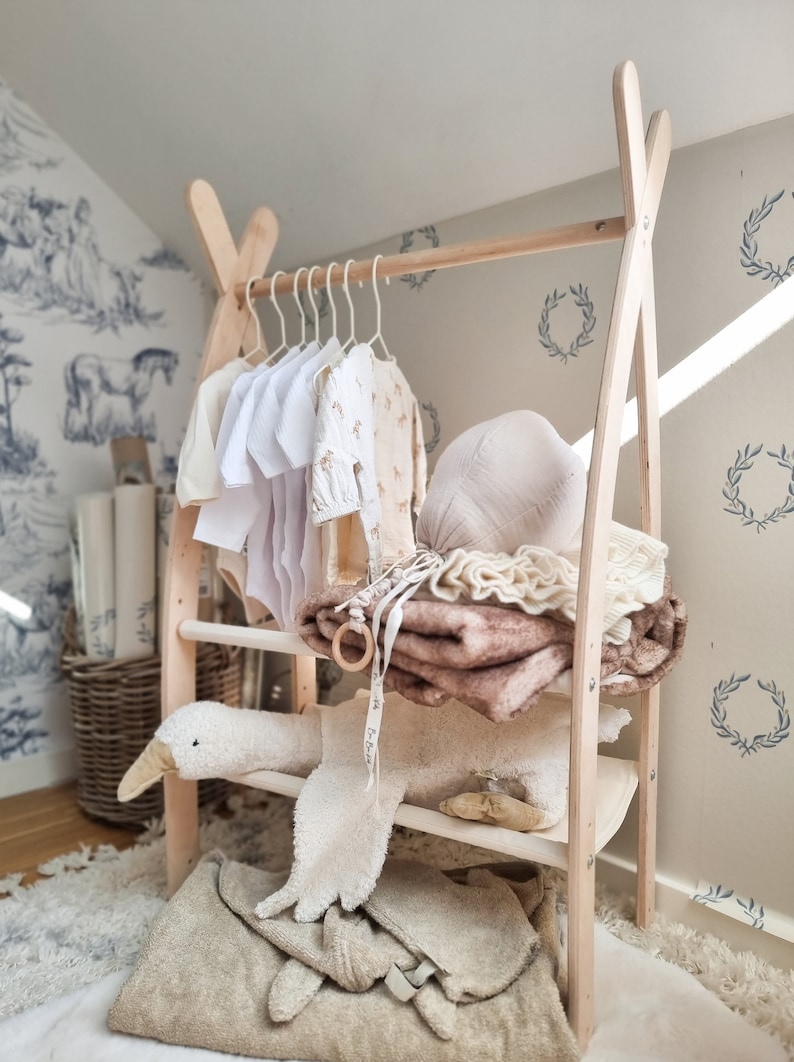 May include: A wooden clothes rack with a white and brown theme. The rack has three shelves and is decorated with a stuffed goose and a variety of baby clothes and blankets.