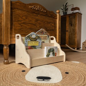 May include: A light-colored wooden floor bookshelf with two tiers for displaying books. The shelf features cut-out handles on the sides and holds three children's books, including titles titled Animal Homes and The Very Hungry Caterpillar.