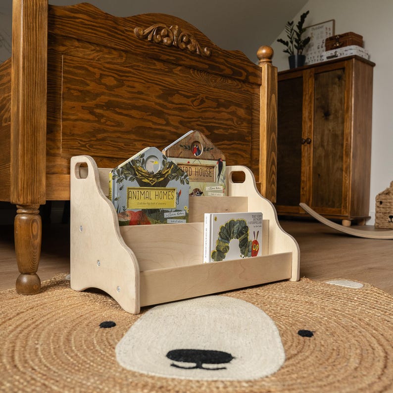 May include: A light-colored wooden floor bookshelf with two tiers for displaying books. The shelf features cut-out handles on the sides and holds three children's books, including titles titled Animal Homes and The Very Hungry Caterpillar.
