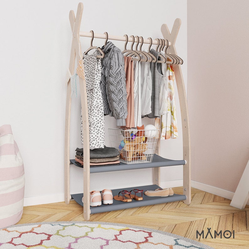 May include: A wooden clothing rack with two shelves and a hanging bar. The rack is made of natural wood and has a light brown finish. The shelves are gray and the hanging bar is made of metal. The rack is decorated with a variety of clothing items, including a sweater, a dress, and a shirt. There is also a small basket on the top shelf, filled with toys. The rack is standing on a wooden floor with a colorful rug in front of it. The text "MAXMOI" is visible on the rug.