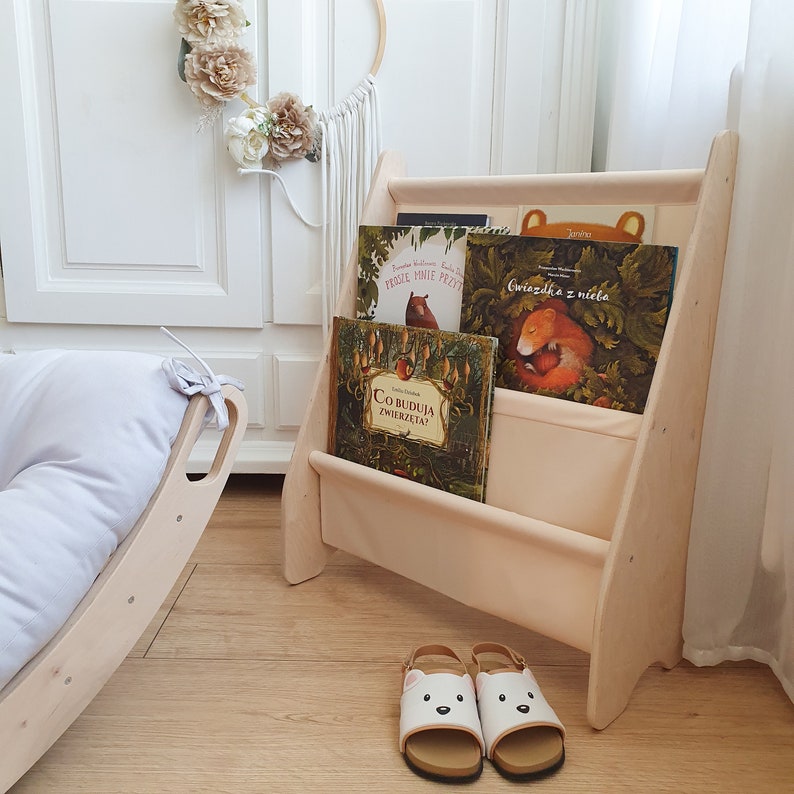 K&ouml;nnte beinhalten: Ein helles Holzregal mit drei Regalbrettern, jedes mit einer Stoffauskleidung. Die Regalbretter sind mit Kinderb&uuml;chern mit bunten Illustrationen gef&uuml;llt. Das Regal steht auf einem Holzboden. Es gibt zwei wei&szlig;e Sandalen mit B&auml;renk&ouml;pfen auf der Vorderseite der Sandalen.