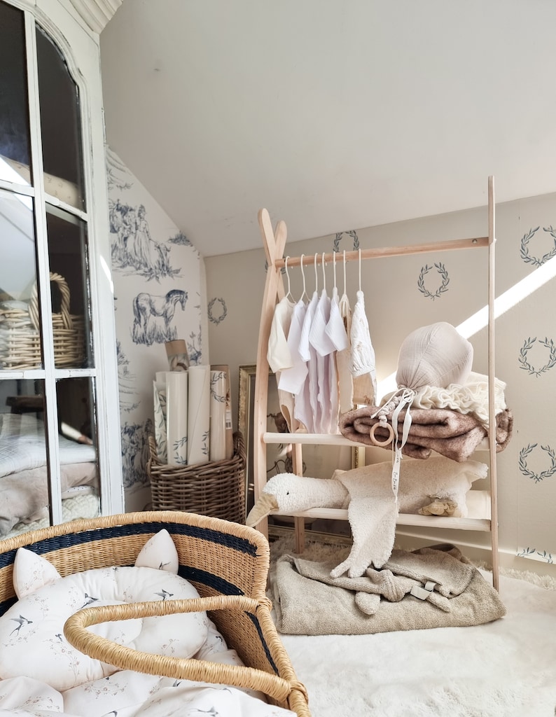 May include: A wooden clothing rack with a white, brown, and beige baby outfit hanging on it. The rack is in a nursery with a wicker basket and a white crib with a floral patterned mattress.