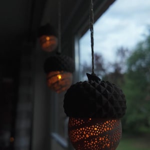 Peut inclure: Trois lampes suspendues en forme de gland avec une lueur chaleureuse. Le dessus de chaque lampe est brun foncé avec un motif texturé, tandis que le bas est brun clair avec un design perforé. Les lampes sont suspendues devant une fenêtre.