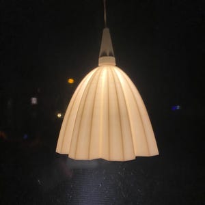 Könnte beinhalten: Eine hängende Pendelleuchte mit cremefarbenem, geripptem Lampenschirm. Die Leuchte hat einen konischen Aufsatz und ein langes, dünnes Kabel. Der dunkle Hintergrund betont den beleuchteten Schirm.