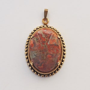 May include: A gold-toned pendant with a red, brown, and gray agate stone set in a beaded oval frame.