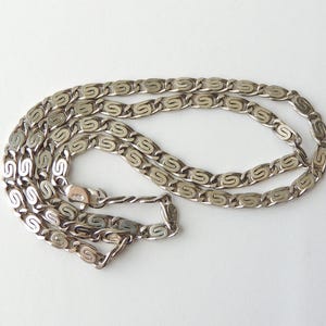 May include: A silver-colored chain necklace with a repeating S-shaped link design. The necklace is laid out on a white surface, showcasing its intricate pattern and the gleam of the metal. The clasp is visible, adding detail to the jewelry.