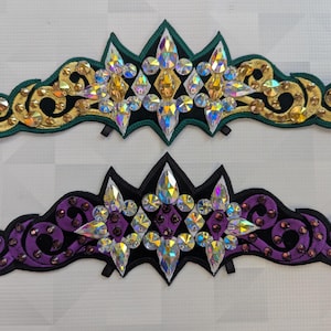 Op de afbeelding: Twee decoratieve tiara's. Eén is groen en goud, de andere is paars en zwart. Beide hebben een centraal stervormig ontwerp met iriserende strass-steentjes en decoratieve krullen. De tiara's zijn waarschijnlijk voor kostuums of speciale evenementen.