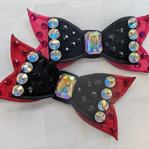 May include: Two decorative hair bows. Each bow has a black centre decorated with iridescent and black rhinestones. The bows have red and pink accents, with additional iridescent rhinestones along the edges. The bows are designed with a butterfly shape.
