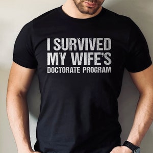 May include: A black t-shirt with white text that reads "I survived my wife's doctorate program".