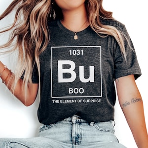 May include: A dark gray t-shirt with a white graphic of a periodic table element. The element is labeled "Bu" with the atomic number "1031" and the name "BOO" below. The text "THE ELEMENT OF SURPRISE" is below the name.