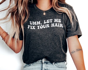 May include: A woman wearing a dark gray t-shirt with the text "Umm, Let Me Fix Your Hair" in white letters. She is wearing light blue jeans and white sneakers. She is sitting on a wooden stool.