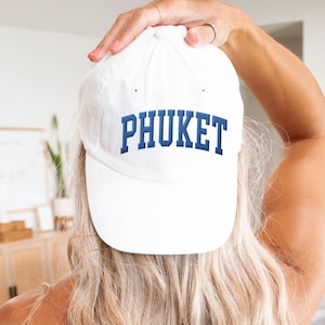 May include: White baseball cap with the word "PHUKET" embroidered in blue block letters. The cap is being held up by a person with long blonde hair. The person is wearing a gray top.