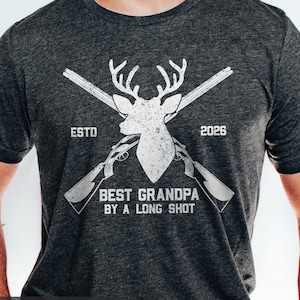 May include: Dark grey heather t-shirt with a white graphic. The graphic features a deer head, crossed shotguns, and the text "BEST GRANDPA BY A LONG SHOT" with "ESTD 2026".