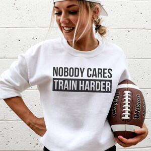 Pu&ograve; includere: Felpa bianca con il testo "Nobody Cares Train Harder" in lettere nere. Una persona tiene un pallone da football marrone e bianco.