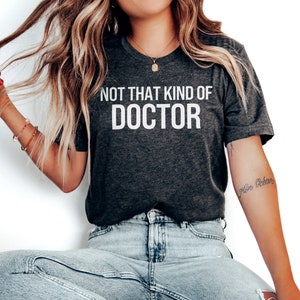 May include: A dark gray t-shirt with white text that reads "Not That Kind Of Doctor". The model is wearing light blue jeans and white sneakers.