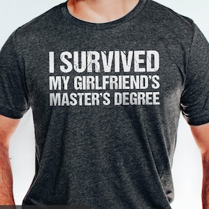 May include: Dark grey heather t-shirt with the white text "I SURVIVED MY GIRLFRIEND'S MASTER'S DEGREE". The words "DARK GREY HEATHER" are at the bottom of the shirt. The shirt is a crew neck and has short sleeves.