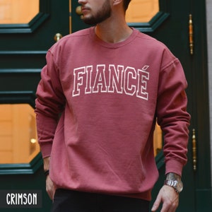 May include: A crimson sweatshirt with the word "FIANCÉ" printed in white on the front.