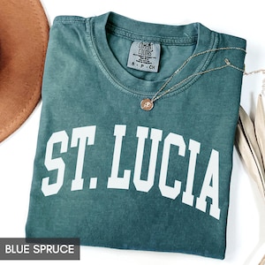 May include: A blue spruce t-shirt with the text "ST. LUCIA" in white block letters. The shirt is folded and has a gold necklace with a pendant. The shirt is made by Comfort Colors.