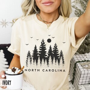 以下が含まれることがあります： クリーム色のTシャツに、満月と「NORTH CAROLINA」の文字が入った黒いシルエットの森林が描かれています。