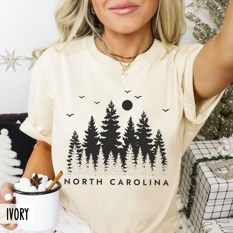 以下が含まれることがあります： クリーム色のTシャツに、満月と「NORTH CAROLINA」の文字が入った黒いシルエットの森林が描かれています。