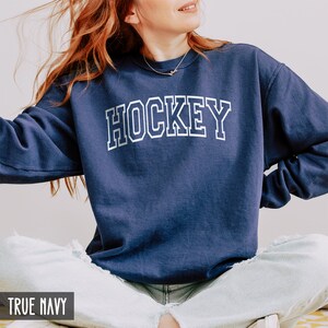 Puede incluir: Una sudadera azul marino con la palabra "HOCKEY" impresa en blanco en la parte delantera. La sudadera est&aacute; hecha de un material suave y c&oacute;modo y tiene un corte holgado.