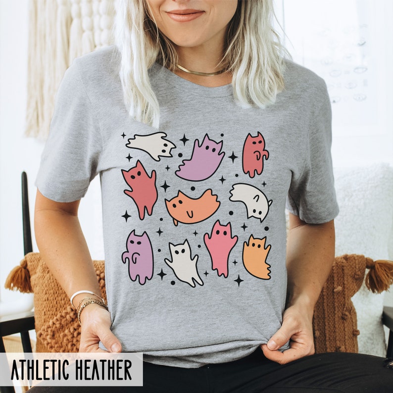Op de afbeelding: Een heidegrijs T-shirt met een grafische print van kleurrijke spookkatten. De katten staan in verschillende poses en hebben verschillende gekleurde ogen. De achtergrond is lichtgrijs met kleine witte sterren.