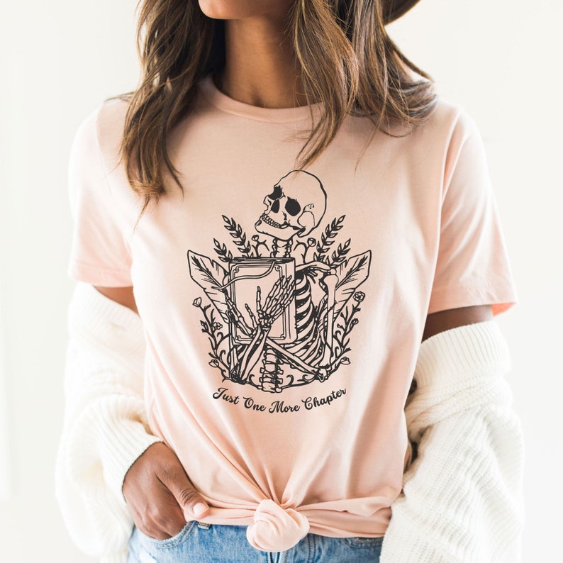May include: A pink t-shirt with a black and white illustration of a skeleton holding a book. The skeleton is surrounded by flowers and leaves. The text "Just One More Chapter" is printed below the illustration.